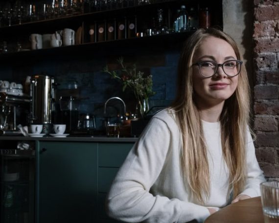 A smiling young woman with eyeglasses sitting in a stylish coffee shop interior.