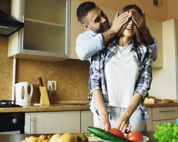 Young man covering girlfriends eyes with hands and surprising her in the kitchen at home. Loving couple cooking breakfast in the morning
